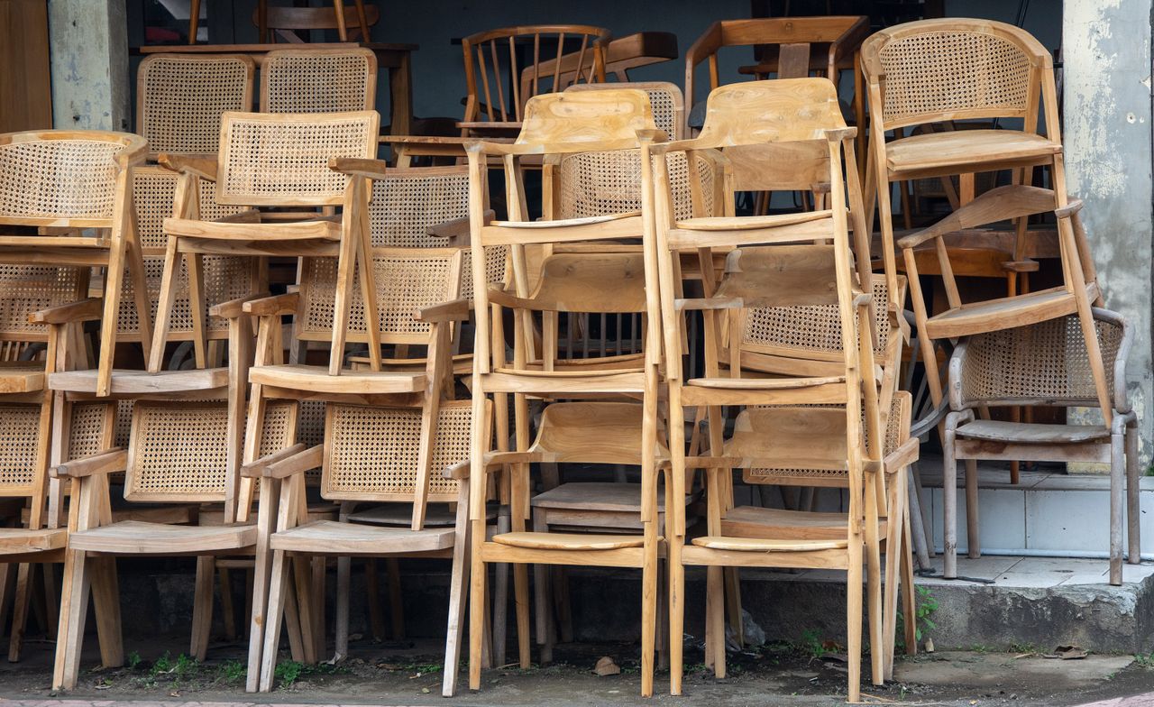 our-experience An array of stacked wooden and wicker chairs leaning against a weathered urban wall.